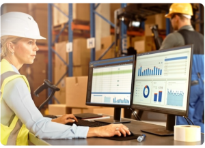 A woman wearing a hi-vis vest and a hard hat sitting at a computer in a warehouse looking at reports